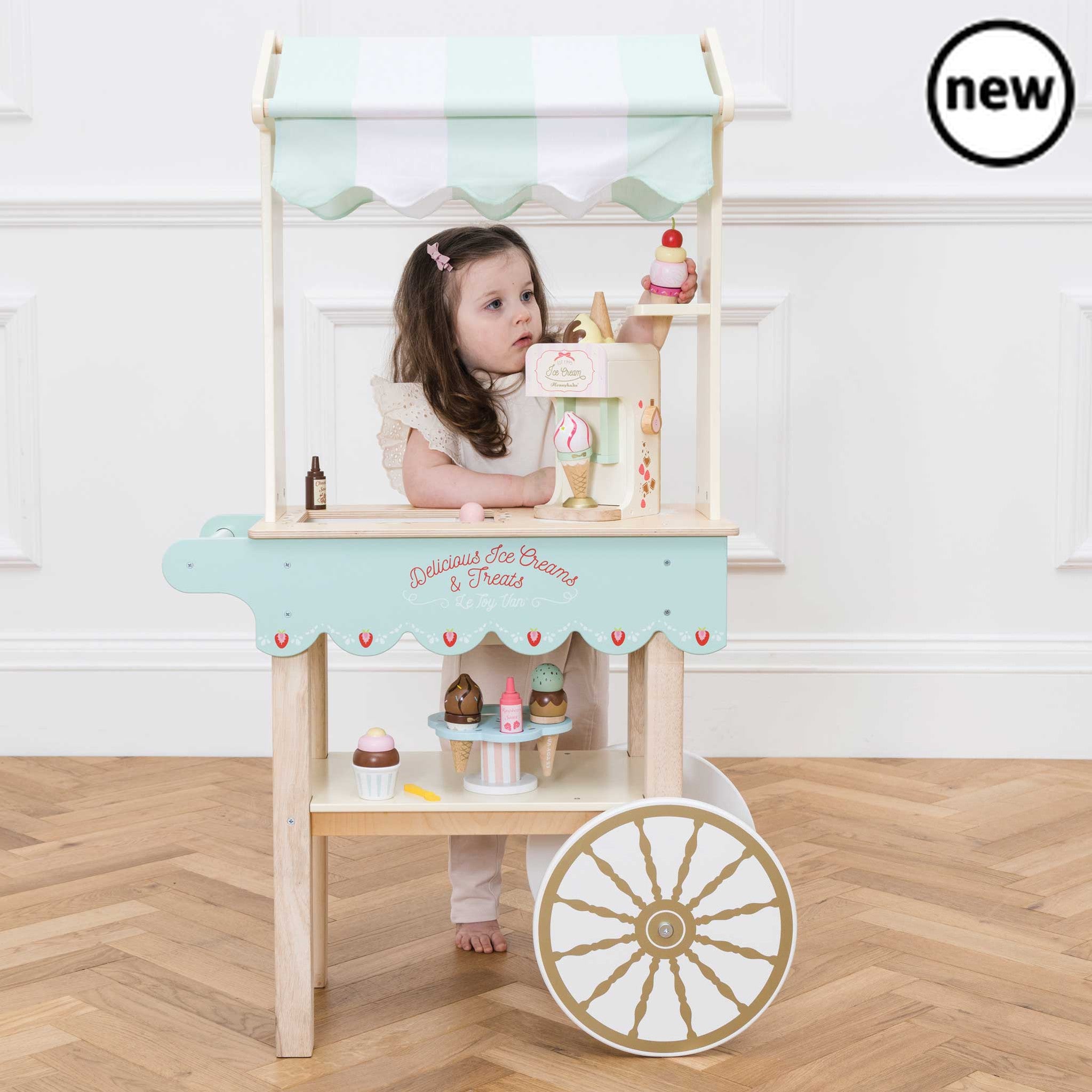 Ice Cream Trolley and Treats, This whimsical Ice-Cream Trolley toy is a nostalgic nod to bygone days and fairs, encouraging both imaginative play and a sense of wonder in children. Key Features Design: The trolley features a barrow style with a classic striped fabric canopy that adds to its vintage allure. Material: Crafted from sustainable wood, this toy is built to last for a lifetime of inspired play. Realistic Features: With an ice compartment featuring a sliding door, the trolley reveals illustrations 
